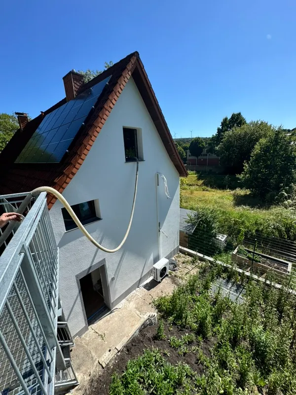 Einblasdämmung an einem Wohnhaus: ein Schlauch führt vom Gerüst zur Dachschräge, unten an der Fassade steht eine Luft-Wärmepumpe